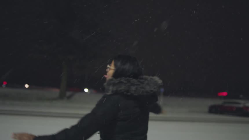 Slow motion footage shows a happy young woman catching snowflakes on her tongue and spinning around outside on a snowy night