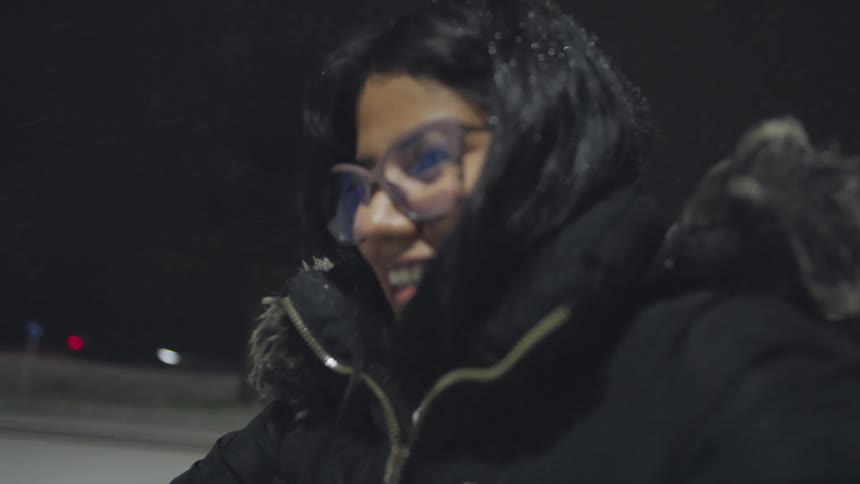 Slow motion shot of a smiling young woman in a cozy winter coat enjoying the falling snow at night