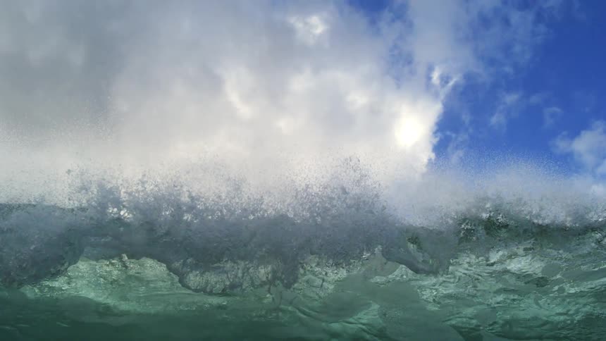 A massive ocean wave crests and breaks in dramatic super slow motion against a partly cloudy blue sky, ending with an underwater view