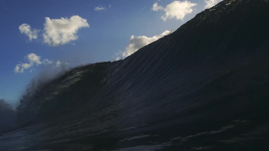 A majestic ocean wave builds and crests in dramatic slow motion, silhouetted against a sunny blue sky before crashing over the camera