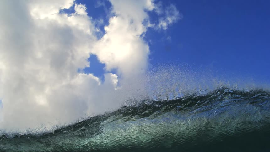 An ocean wave crests and breaks in dramatic slow motion, revealing the turbulent beauty of the water against a blue sky with clouds before plunging underwater