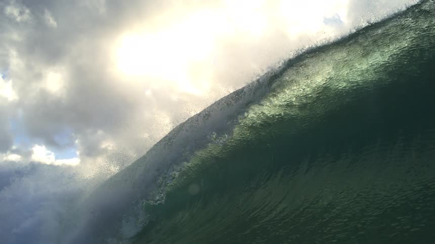 A super slow-motion view from inside a massive, curling ocean wave as the sun breaks through the clouds