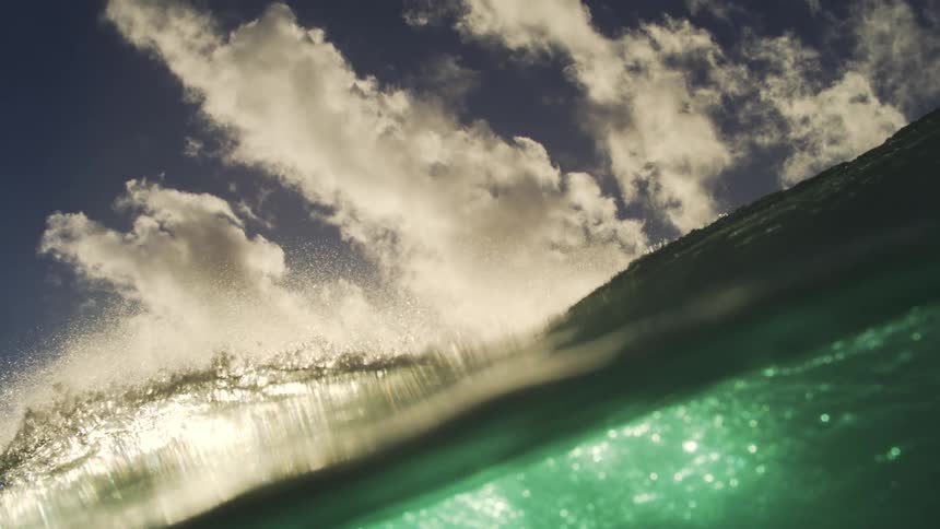 A slow-motion, immersive view from the ocean as a large, dark wave rises and breaks over the camera, transitioning to an underwater perspective with shimmering light