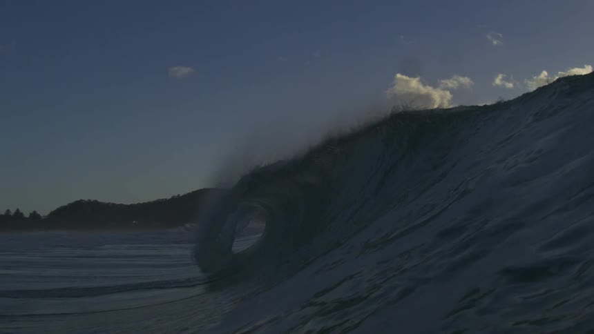 An epic slow-motion view captures a massive ocean wave forming a perfect barrel and breaking against the morning sky