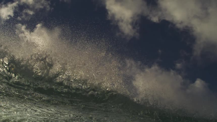 A powerful ocean wave crests and breaks in extreme slow motion, filmed from a low angle against a dramatic cloudy sky