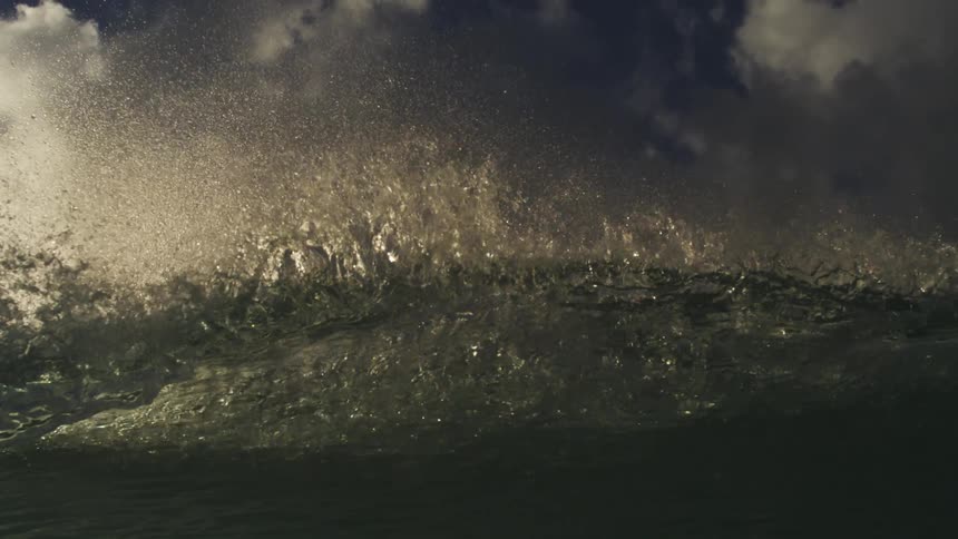 Dramatic slow motion close-up of a powerful ocean wave's backlit spray against a dark, stormy sky before submerging underwater
