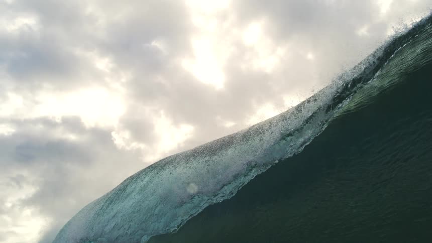 An immense ocean wave builds and breaks in spectacular slow motion, shot from water level against a dramatic cloudy sky