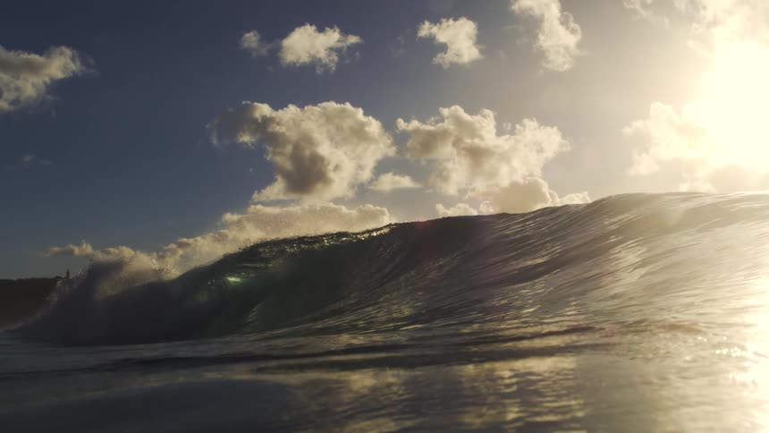 An epic ocean wave crests in super slow motion, silhouetted against a bright sun and cloudy sky