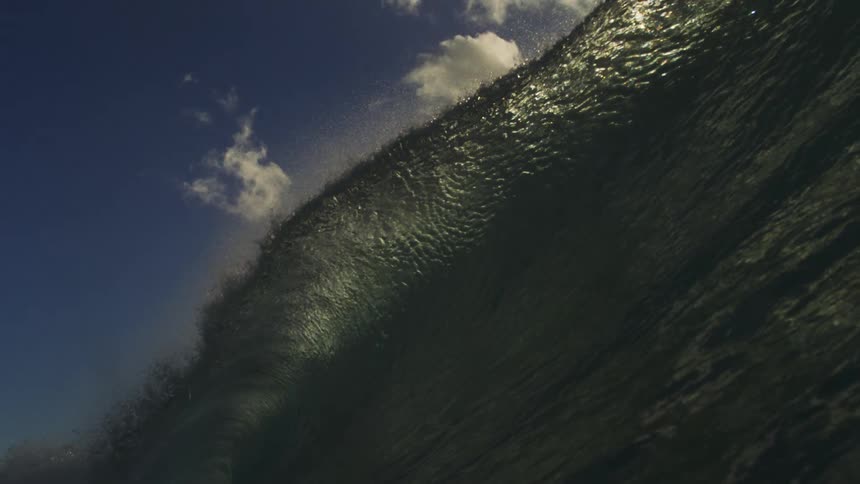 An immense ocean wave crests in dramatic slow motion, its surface shimmering as it's backlit by the sun against a partly cloudy sky