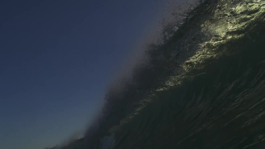 A massive ocean wave crests in dramatic super slow motion against a beautiful blue sky
