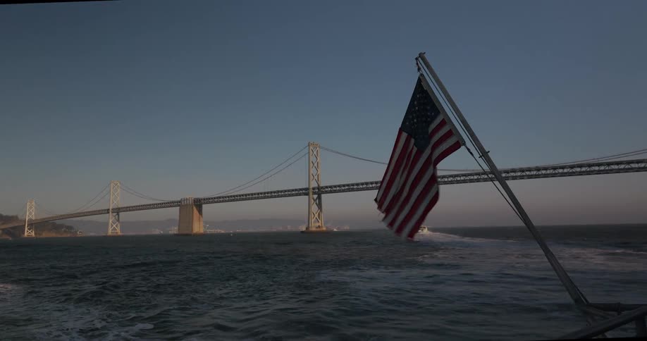 Slow motion view of an American flag waving from a boat with the San Francisco-Oakland Bay Bridge in the background at sunset