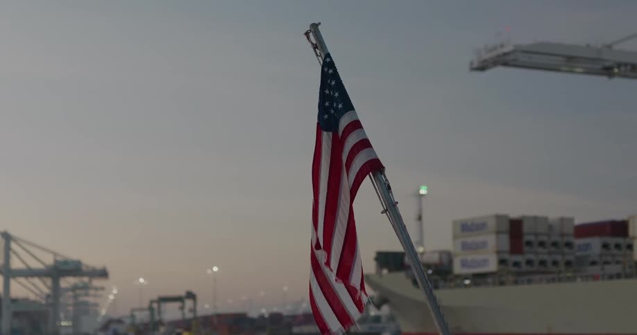 A still American flag hangs over a busy industrial shipping port with a large cargo container ship in the background at twilight