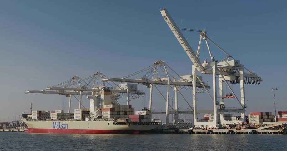 A large Matson container ship is docked at an industrial port while gantry cranes load and unload cargo under a clear blue sky