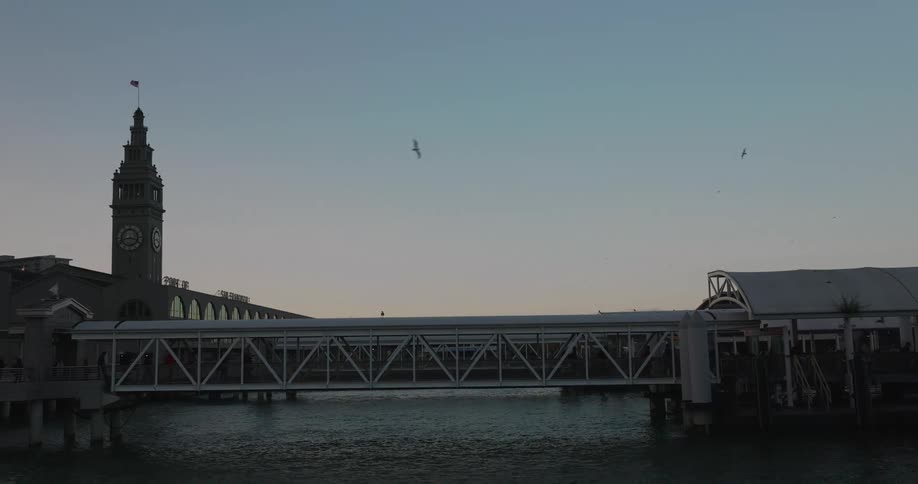 A tranquil, static shot captures the San Francisco Ferry Building at dusk as people walk along the pier's gangway over the calm bay