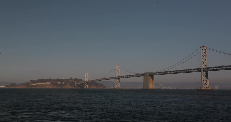 A static wide shot shows the San Francisco-Oakland Bay Bridge spanning across the bay on a clear, sunny day