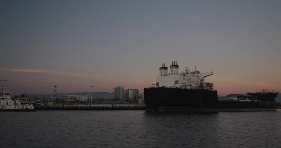A massive industrial cargo ship is docked in a calm harbor at dusk with a city skyline in the background