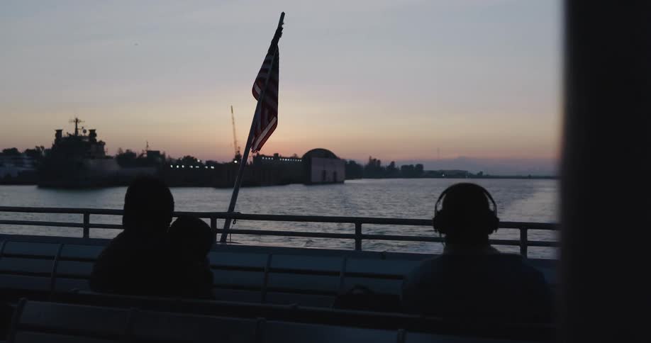 Passengers relax on a ferry at sunset, with an American flag waving over the water as they depart from an industrial port