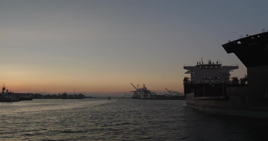 A massive cargo ship is docked in an industrial harbor as the sun sets over the water, with cranes silhouetted in the distance