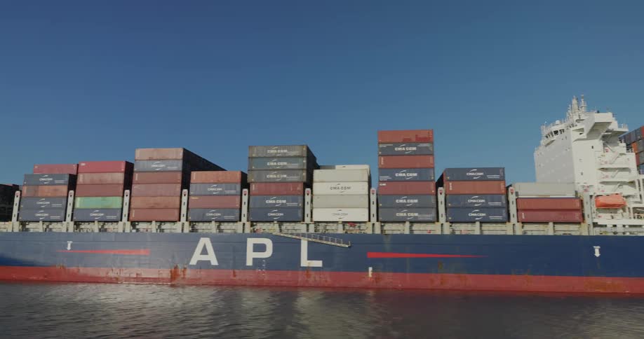 Slow pan alongside a huge container cargo ship moored at an industrial shipping port under a clear blue sky