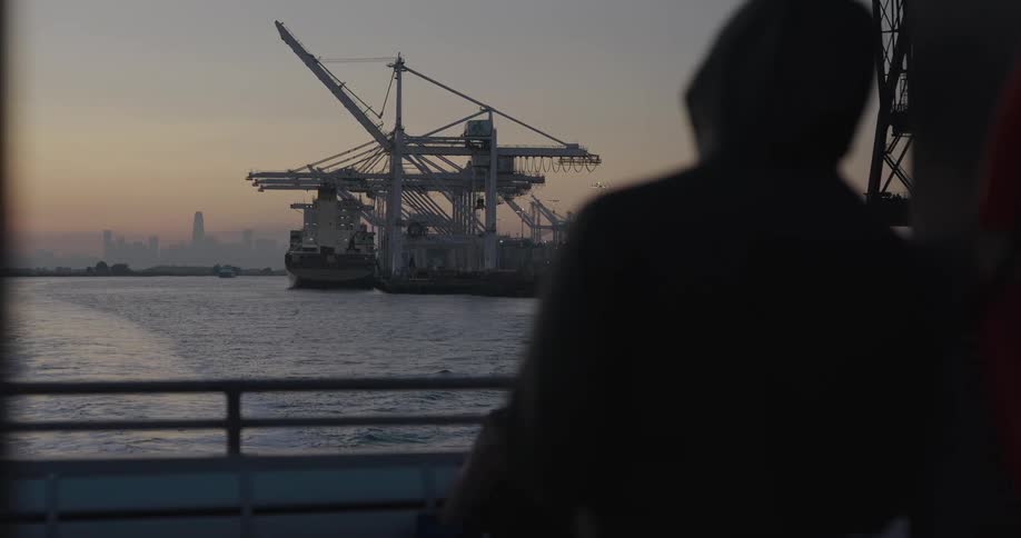 A silhouetted person looks out from a boat at a large industrial shipping port with cranes at sunset, as a distant city skyline is visible on the horizon