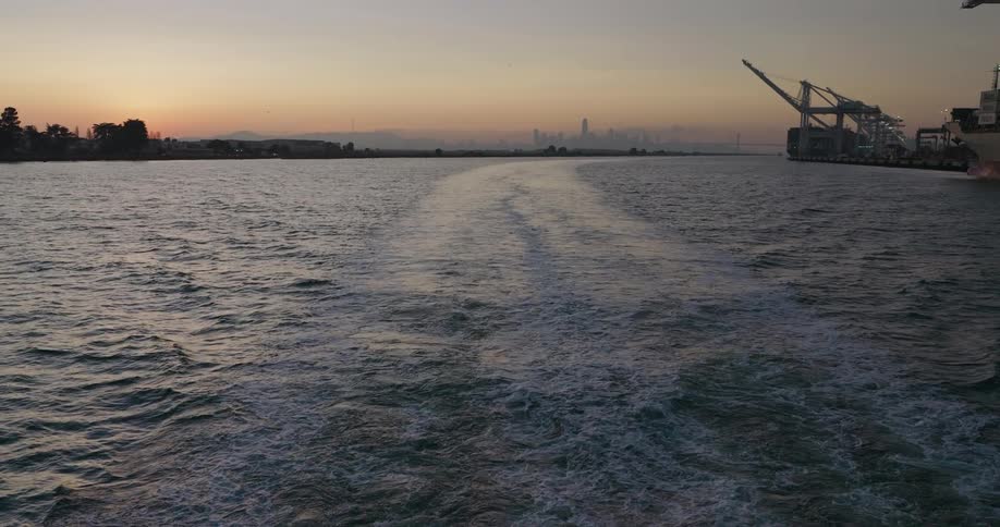A view of the wake from the back of a boat at sunset in an industrial harbor, with a city skyline in the distant haze