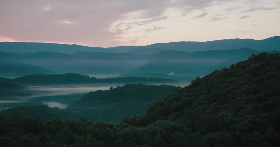 Drone shot pulling back from a forested hill to reveal a vast, foggy mountain valley at dawn