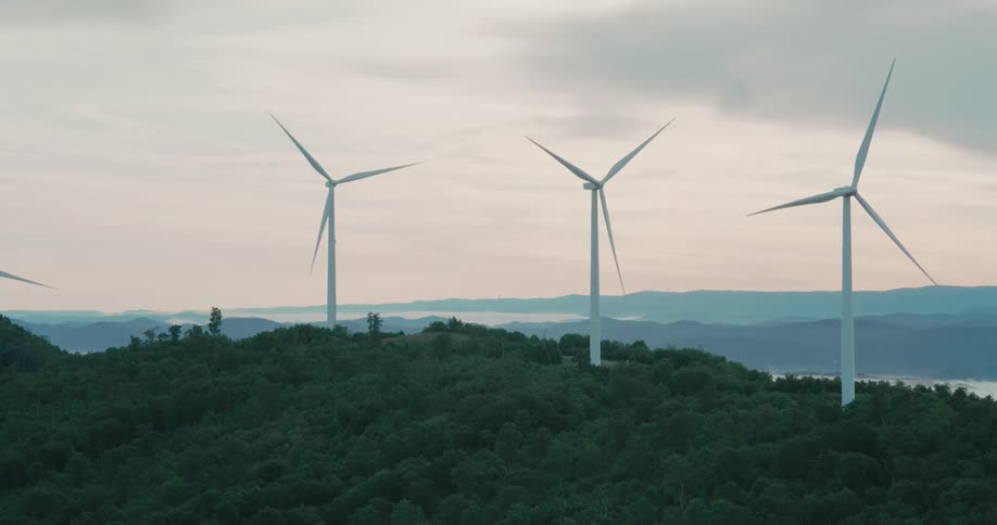 Large wind turbines spin slowly on a lush green mountain top with a hazy, mountainous landscape in the background