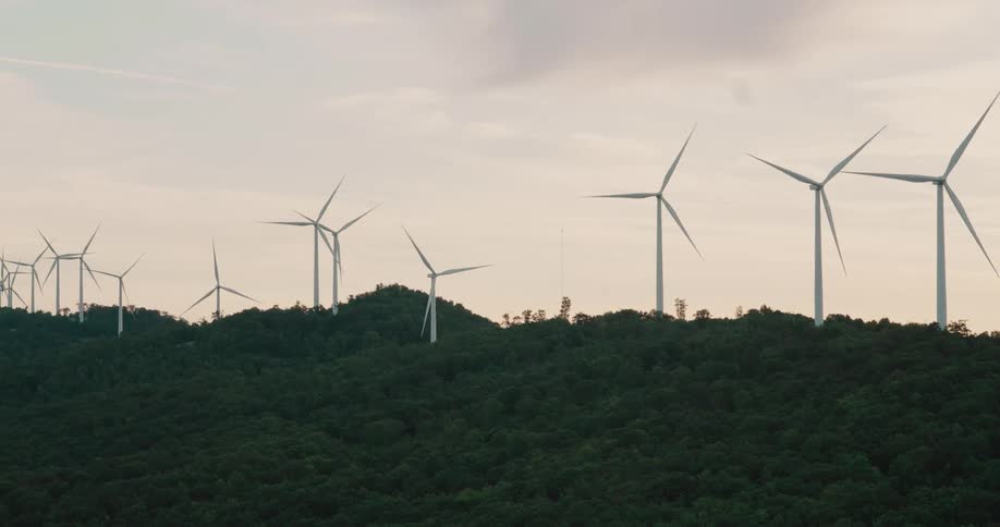 A wind farm with multiple turbines spins on a lush green forested hilltop under a cloudy sky