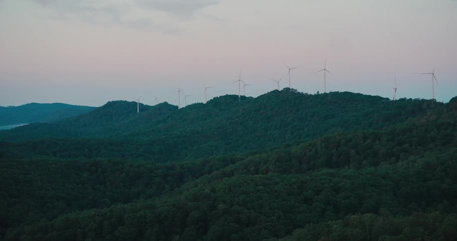 A wide shot of spinning wind turbines atop lush, green hills at dusk
