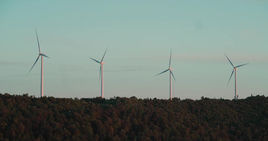 A slow panoramic view of multiple wind turbines spinning on a forested mountain top at sunset