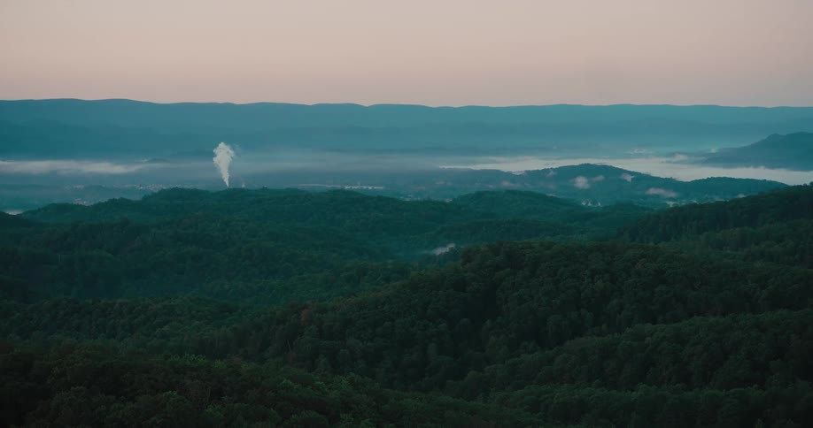 A tranquil, wide view of rolling green mountain forests at dawn with a layer of fog in the valley and a single smokestack in the distance