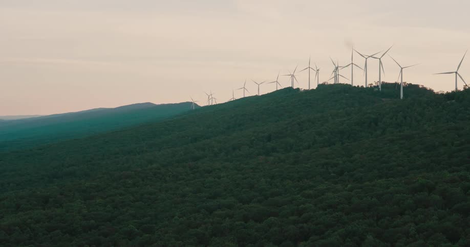 A row of wind turbines spins atop a lush, green mountain ridge under an overcast sky