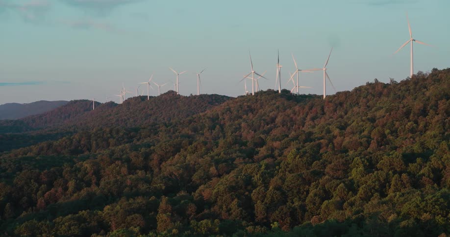 A row of modern wind turbines spins atop a lush, forested mountain range during a serene sunrise