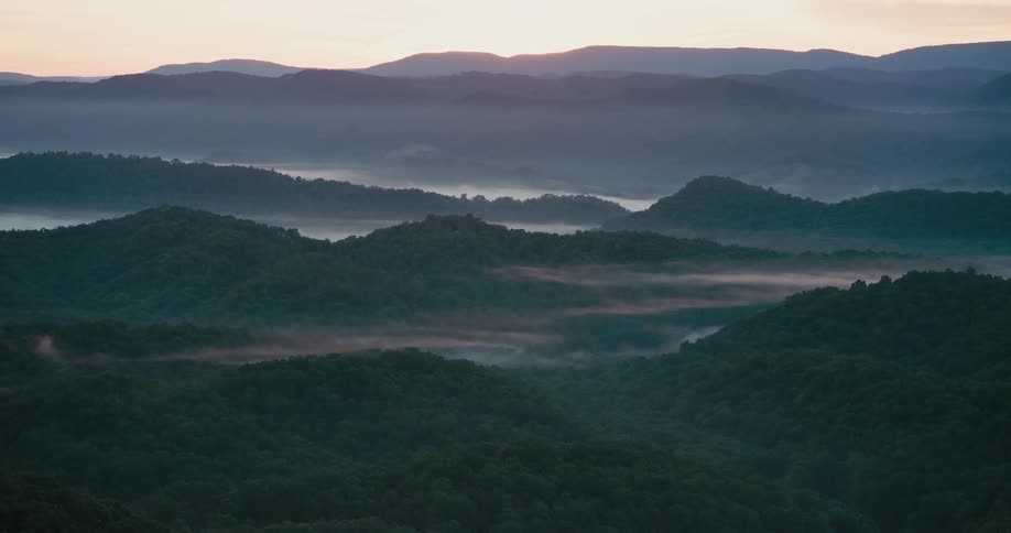 A tranquil sunrise over a vast mountain range with thick fog settling in the forest-covered valleys