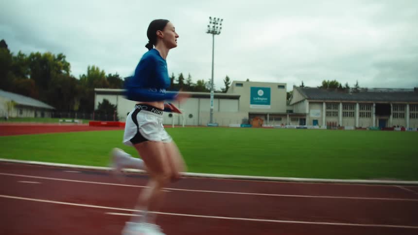 A determined female athlete sprints with focus and intensity on a red running track at an outdoor stadium