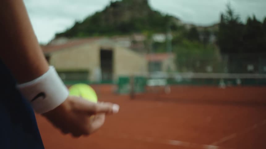 A female tennis player in slow motion tosses a tennis ball in her hand while standing on a clay court