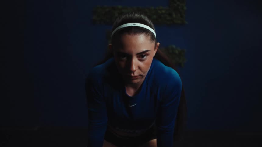 A determined female athlete in a dark gym looks up and stares intensely into the camera