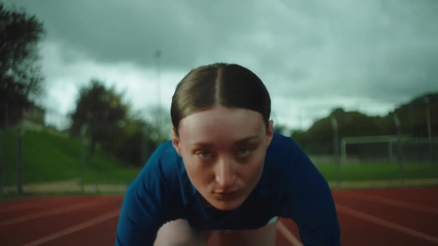 A determined female sprinter looks up with intense focus from the starting blocks on a running track