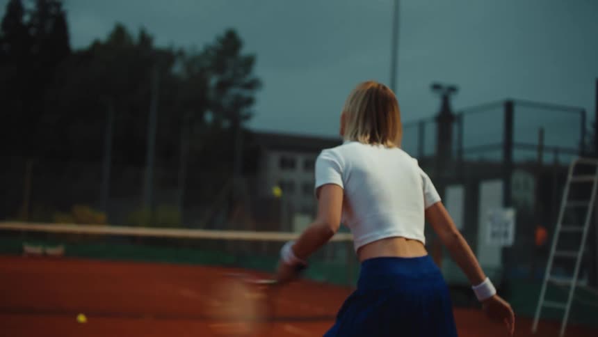 A female tennis player hits forehand shots in slow motion on a clay court during a moody evening practice session