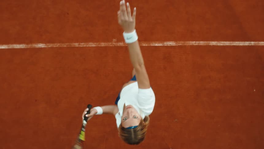 A female tennis player serves the ball in a powerful top-down slow-motion shot on a red clay court