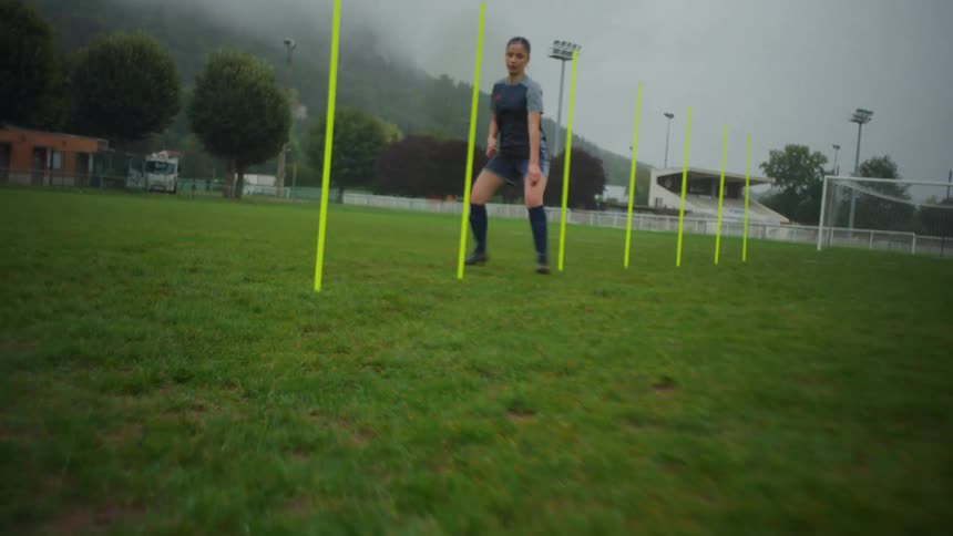 Slow motion tracking shot of a female soccer player training her agility by weaving through yellow poles on an overcast day