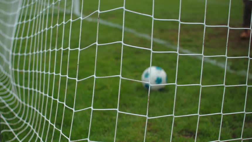 A soccer ball flies into the back of the net in slow motion, scoring a goal on a green field