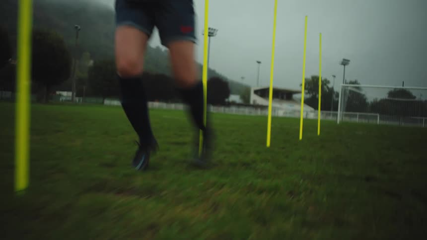 A determined female football player trains on a foggy field, weaving through yellow agility poles in slow motion