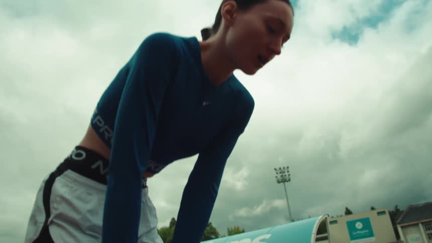 A female athlete in athletic wear catches her breath on a running track under an overcast sky