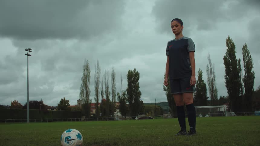 A focused female soccer player practices her penalty kick on an empty field under a dramatic cloudy sky