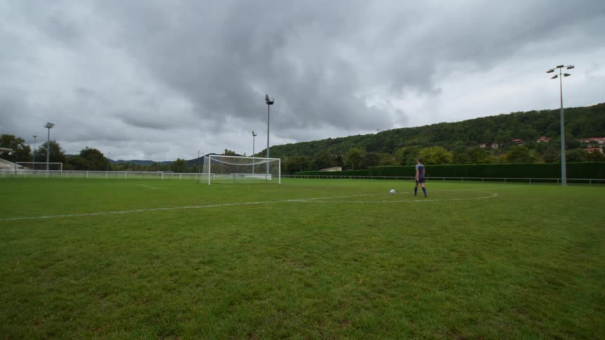 A solitary soccer player sets up a ball for a kick on an empty field under an overcast sky