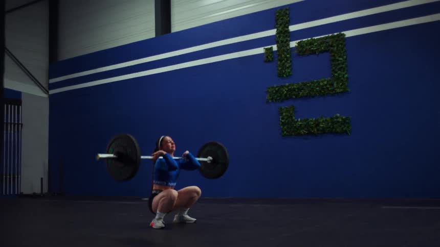 A young female weightlifter fails a heavy barbell clean, losing her balance and falling backward in slow motion at the gym