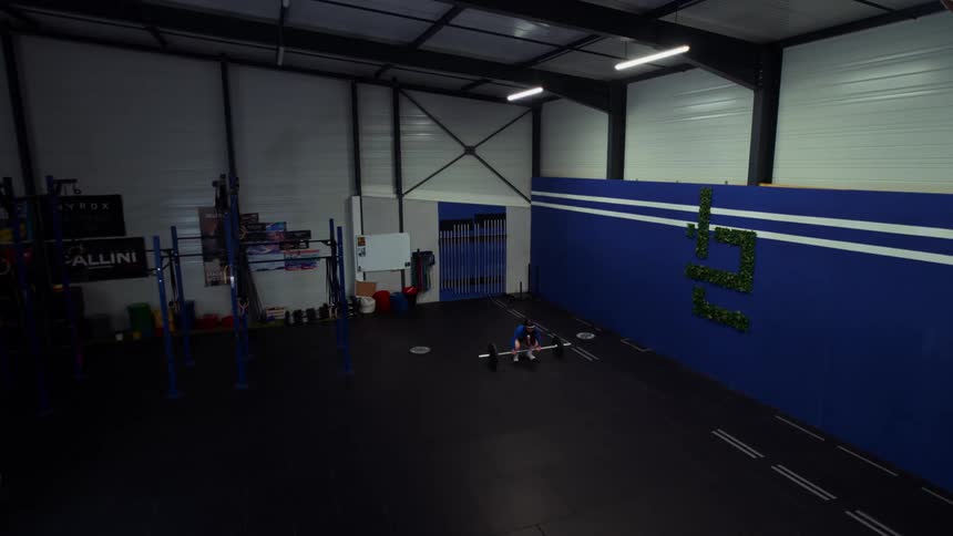 A lone male athlete performs a powerful clean lift with a barbell in a vast, empty gym, viewed from a high angle