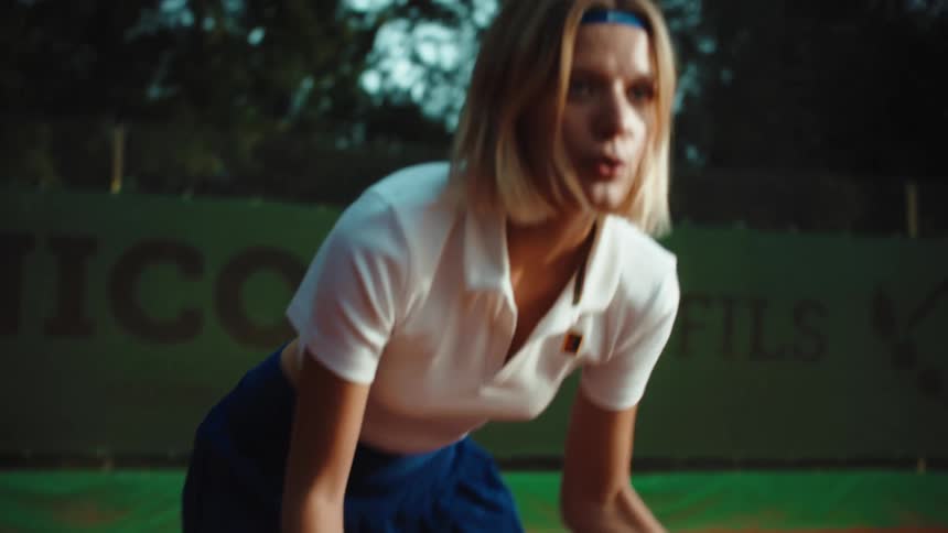A determined female tennis player bounces a ball on her racket before tilting up to reveal her focused expression on a clay court