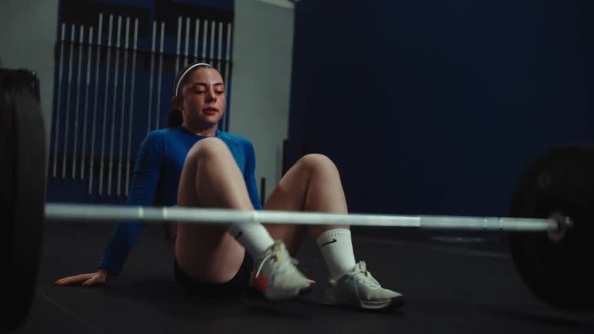 A female weightlifter in a gym strains to lift a heavy barbell but fails, dropping it and sitting on the floor to catch her breath in slow motion